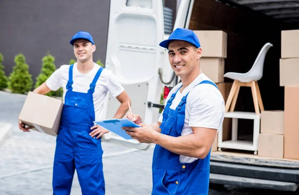 Two professional movers in blue uniforms and caps smile at the camera while loading or unloading boxes and furniture from a moving truck.