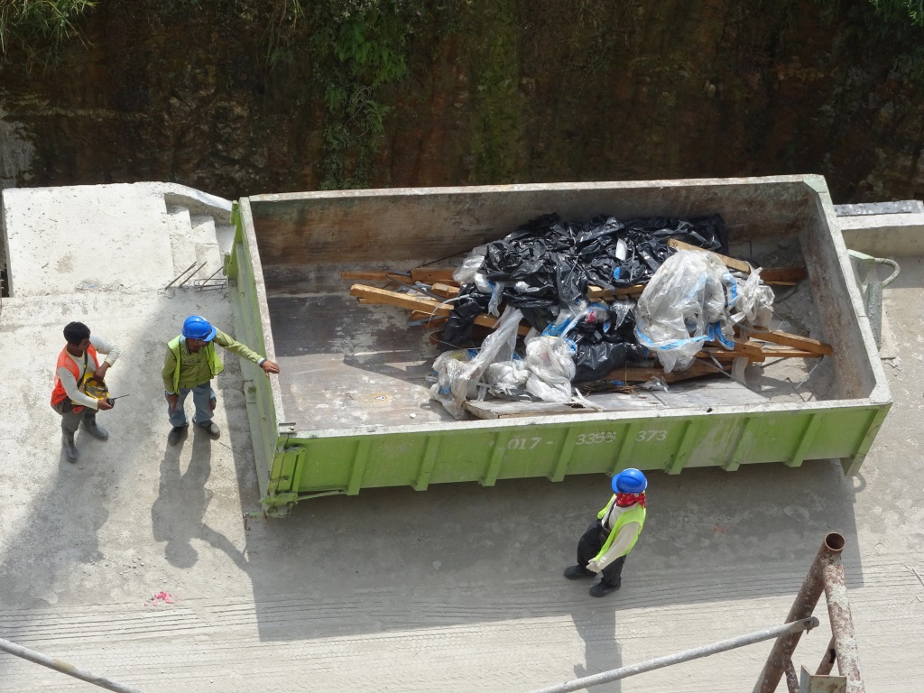 Three construction workers in high-visibility safety gear stand around a large green dumpster filled with construction debris and trash bags on a concrete surface.