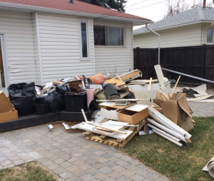 A large pile of household debris including cardboard boxes, furniture, appliances, and various items sits on the driveway of a residential home, likely from a cleanout or renovation project.RetryClaude can make mistakes. Please double-check responses.